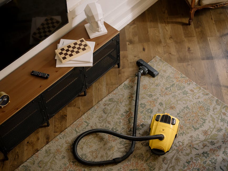 A person kneeling on a patterned carpet in a living room, opening a yellow and black portable vacuum cleaner with a flexible hose attached. The carpet shows signs of wear, with some dirt visible, indi