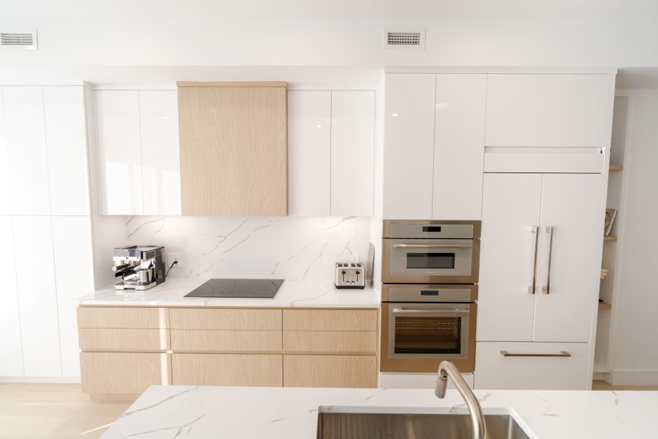 A modern, minimalist kitchen featuring white high-gloss cabinets and a light wood finish on lower drawers. The kitchen has a white marble backsplash with subtle grey veining, complementing the light w