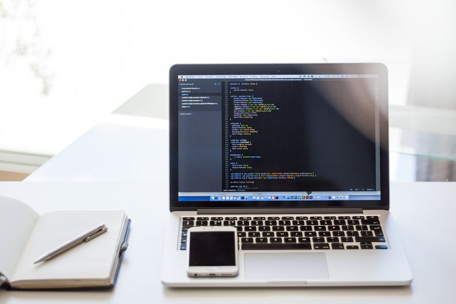 A white desk in a well-lit room, featuring an open laptop displaying lines of code on a dark background, positioned towards the right side of the image. To the left of the laptop, there is a closed no